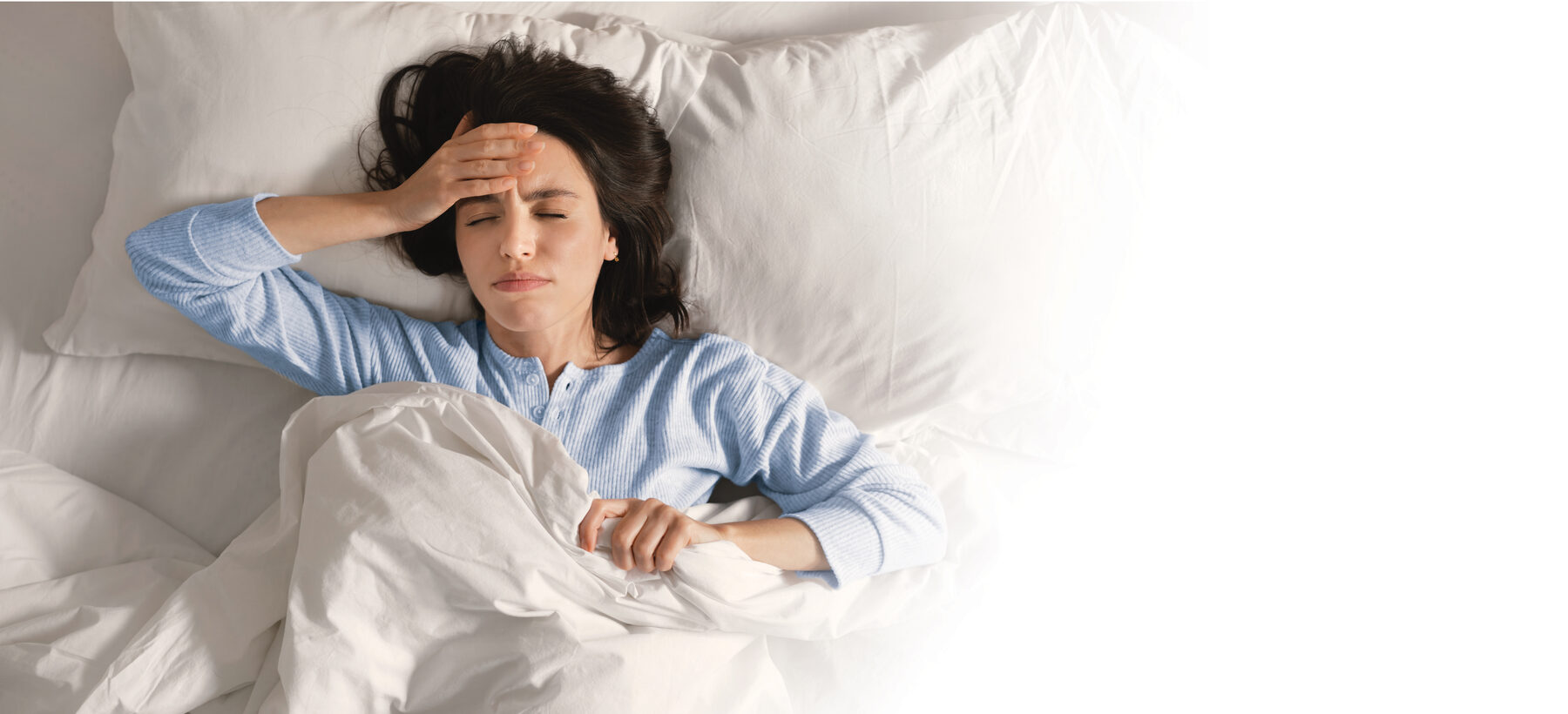 Woman lying in bed experiencing a morning migraine, holding her forehead.